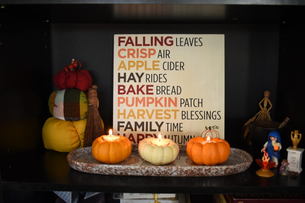 Photo of an altar with pumpkin themed items, a miniature Hestia, a sign about the fall, and candles.